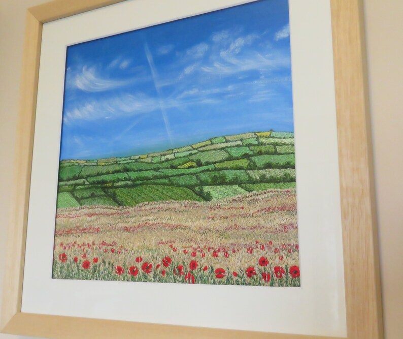 May include: A framed landscape painting depicting a field of red poppies, a wheat field, and green hills under a blue sky with clouds. The artwork is presented in a light wooden frame. The painting evokes a rural scene, with a focus on natural elements.