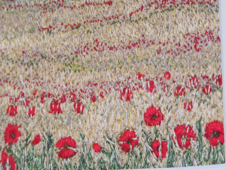 May include: A textile artwork showing a field of wheat and red poppies. The piece uses textured stitching in beige, green, and red. The poppies are a bright red, and the wheat field is a light beige. The artwork is a close-up view.