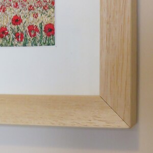May include: A framed print depicting a field of red poppies. The artwork is presented with a white mount and a light-coloured wooden frame. The frame's corner is visible, showing the natural wood grain.