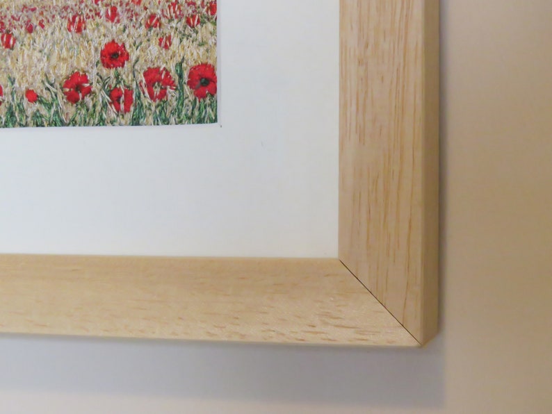 May include: A framed print depicting a field of red poppies. The artwork is presented with a white mount and a light-coloured wooden frame. The frame's corner is visible, showing the natural wood grain.