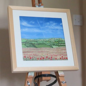 May include: A framed landscape painting depicting a field of red poppies beneath a blue sky with wispy clouds. The artwork, in a light wood frame, is displayed on an easel. The scene includes green rolling hills and fields.