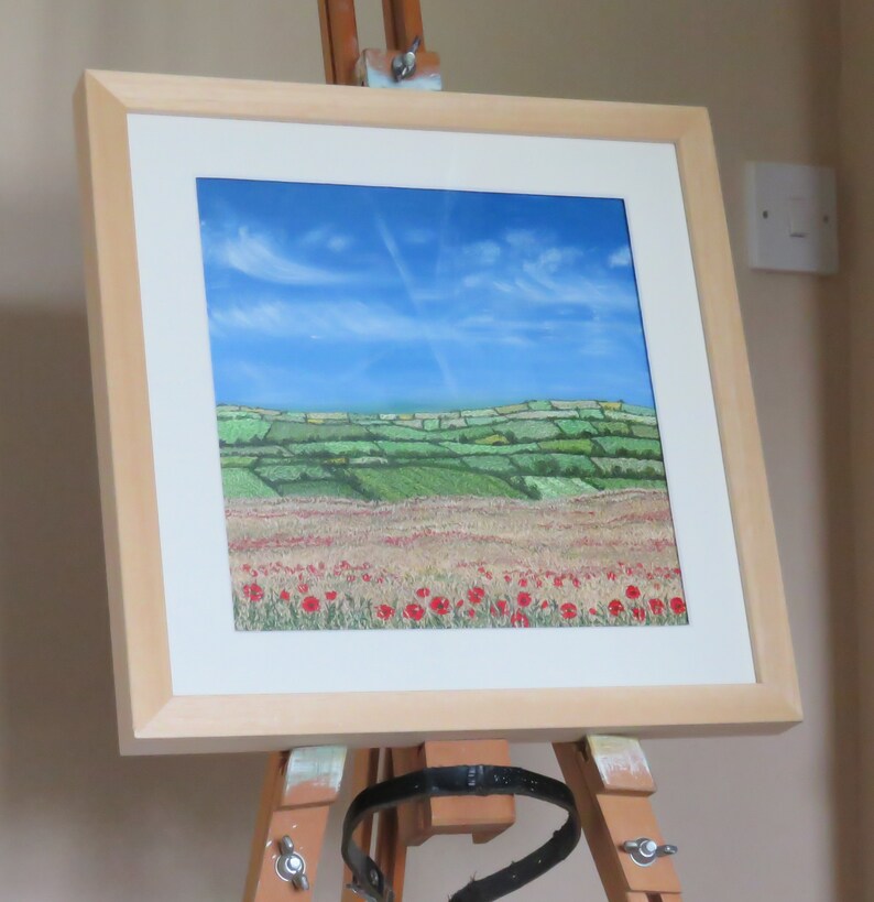 May include: A framed landscape painting depicting a field of red poppies beneath a blue sky with wispy clouds. The artwork, in a light wood frame, is displayed on an easel. The scene includes green rolling hills and fields.