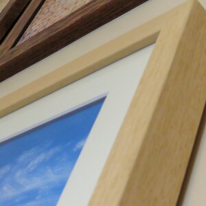 May include: Close-up of framed artwork with a light wood frame and a white mount. The artwork features a blue sky with white clouds. Another framed piece with a dark wood frame is visible.