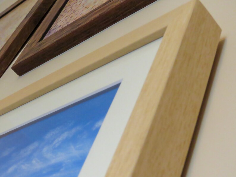 May include: Close-up of framed artwork with a light wood frame and a white mount. The artwork features a blue sky with white clouds. Another framed piece with a dark wood frame is visible.