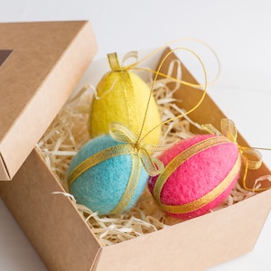 Op de afbeelding: Een set van drie kleurrijke paasei-ornamenten in een bruine kartonnen doos. De eieren zijn geel, blauw en roze, elk versierd met een gouden lint. De doos is gevuld met stro-achtig verpakkingsmateriaal.