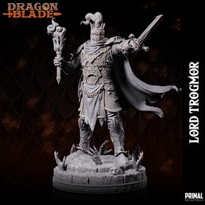 Op de afbeelding: Een grijs geverfd beeldje van een ridder die een zwaard en een toorts vasthoudt. De ridder draagt ​​een harnas en een cape. Het beeldje staat op een voetstuk met een schedel en botten. De tekst "Dragon Blade" en "Lord Trogmor" is zichtbaar op de afbeelding.