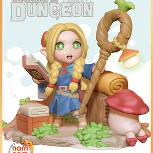 May include: A figurine from "Delicious in Dungeon" depicting a blonde character in a blue outfit, holding a book and staff. The figure is set on a base with a mushroom, a wooden box, and green foliage. The text "Delicious in Dungeon" is at the top.