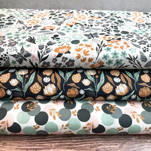 Könnte beinhalten: Drei Stoffrollen mit floralen und geometrischen Mustern. Die obere Rolle hat einen weißen Hintergrund mit orangefarbenen, schwarzen und grauen Blumen und Schmetterlingen. Die mittlere Rolle hat einen dunkelblauen Hintergrund mit tanfarbenen und weißen Blumen. Die untere Rolle hat einen weißen Hintergrund mit grünen und grauen Kreisen.