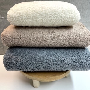May include: Three plush blankets stacked on a wooden stand. The top blanket is white, the middle blanket is beige, and the bottom blanket is gray.