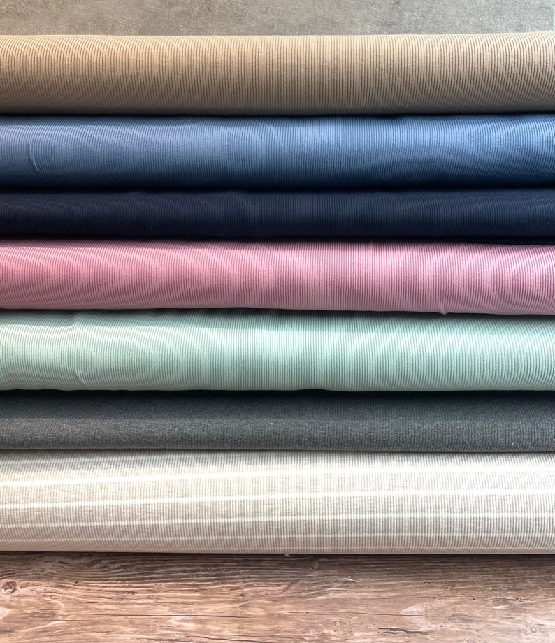 May include: A stack of eight different colored ribbed fabrics. The fabrics are arranged in a vertical stack, with the top fabric being a light brown color. The fabrics are all ribbed and have a soft, textured feel.