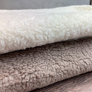 May include: Two pieces of fluffy fabric, one white and one beige, are folded and stacked on top of each other. The fabric has a soft, fuzzy texture.