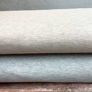 May include: Two rolls of ribbed fabric, one in a light beige color and the other in a light gray color. The fabric is folded and stacked on top of a wooden surface.