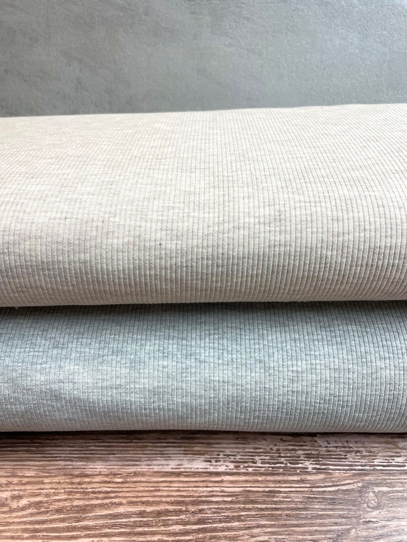 May include: Two rolls of ribbed fabric, one in a light beige color and the other in a light gray color. The fabric is folded and stacked on top of a wooden surface.