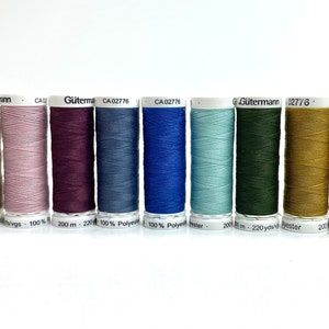 Könnte beinhalten: Eine Reihe von 12 Garnrollen in verschiedenen Farben, darunter Weiß, Beige, Rosa, Lila, Blau, Grün, Braun und Schwarz. Die Rollen sind mit dem Markennamen "Gütermann" und dem Garn-Typ "Polyester" gekennzeichnet.