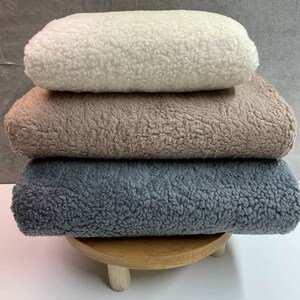May include: Three plush blankets stacked on a wooden stand. The top blanket is white, the middle blanket is brown, and the bottom blanket is gray.