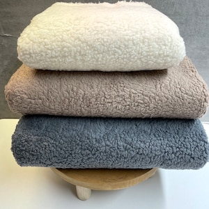 May include: Stack of four plush blankets in white, beige, gray, and dark gray. The blankets are folded and stacked on a wooden pedestal.