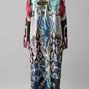 May include: A long-sleeved, floor-length robe featuring a vibrant, multi-colored ikat pattern in shades of blue, green, white, black, and red. The garment has a V-neckline and is adorned with a decorative, dark-toned brooch on the upper chest area.