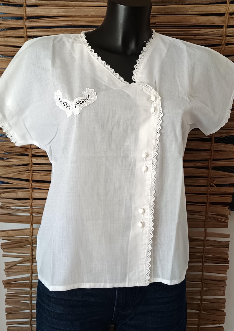 May include: White short-sleeved blouse with delicate lace trim along the neckline, sleeves, and button placket. Features a decorative lace applique on the left side. The blouse has a button-up front and is made of a lightweight fabric.