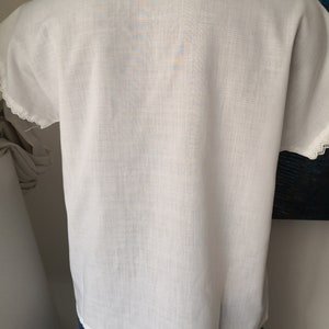 May include: White short-sleeved blouse with a V-neck and delicate lace trim on the sleeves. The fabric appears to be lightweight and slightly textured. The blouse is paired with blue denim jeans.