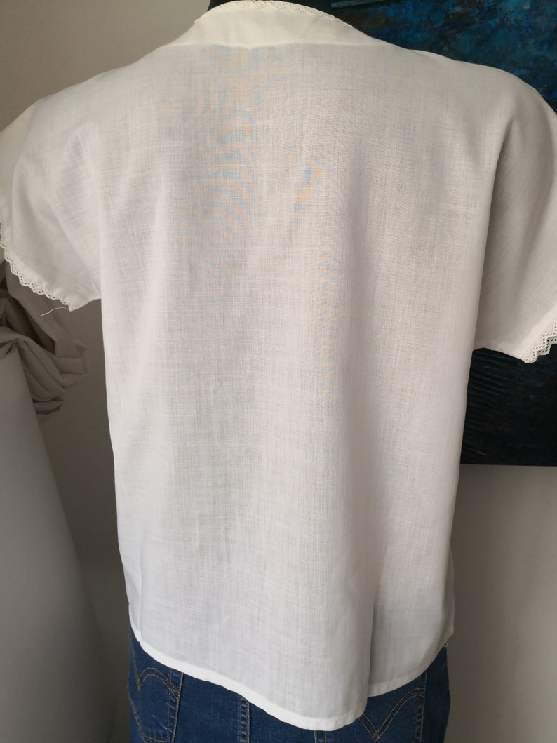 May include: White short-sleeved blouse with a V-neck and delicate lace trim on the sleeves. The fabric appears to be lightweight and slightly textured. The blouse is paired with blue denim jeans.