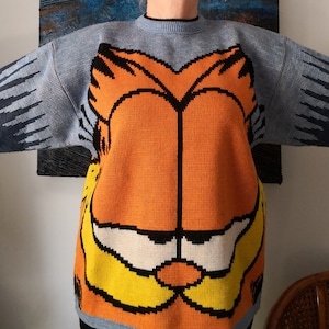 Peut inclure: Un pull bleu clair avec un grand visage orange de Garfield sur le devant. Le pull a des manches longues avec un motif bleu et gris sur le haut des bras. Le col et les poignets sont noirs.
