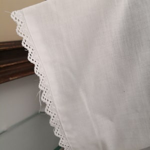 May include: Close-up of a white fabric sleeve with a delicate, scalloped lace trim along the edge. The fabric appears to be a lightweight material, possibly linen or cotton, with a subtle texture. The sleeve is angled, showing the detail of the lace.
