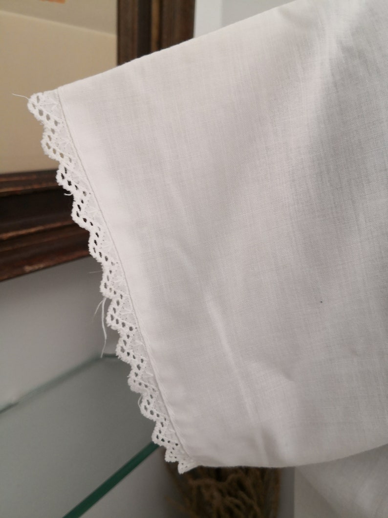 May include: Close-up of a white fabric sleeve with a delicate, scalloped lace trim along the edge. The fabric appears to be a lightweight material, possibly linen or cotton, with a subtle texture. The sleeve is angled, showing the detail of the lace.
