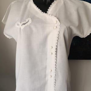 May include: White short-sleeved blouse with a wrap-style front. The blouse features delicate lace trim along the neckline, sleeves, and front closure. A small lace applique is on the left side. The blouse is buttoned down the front.