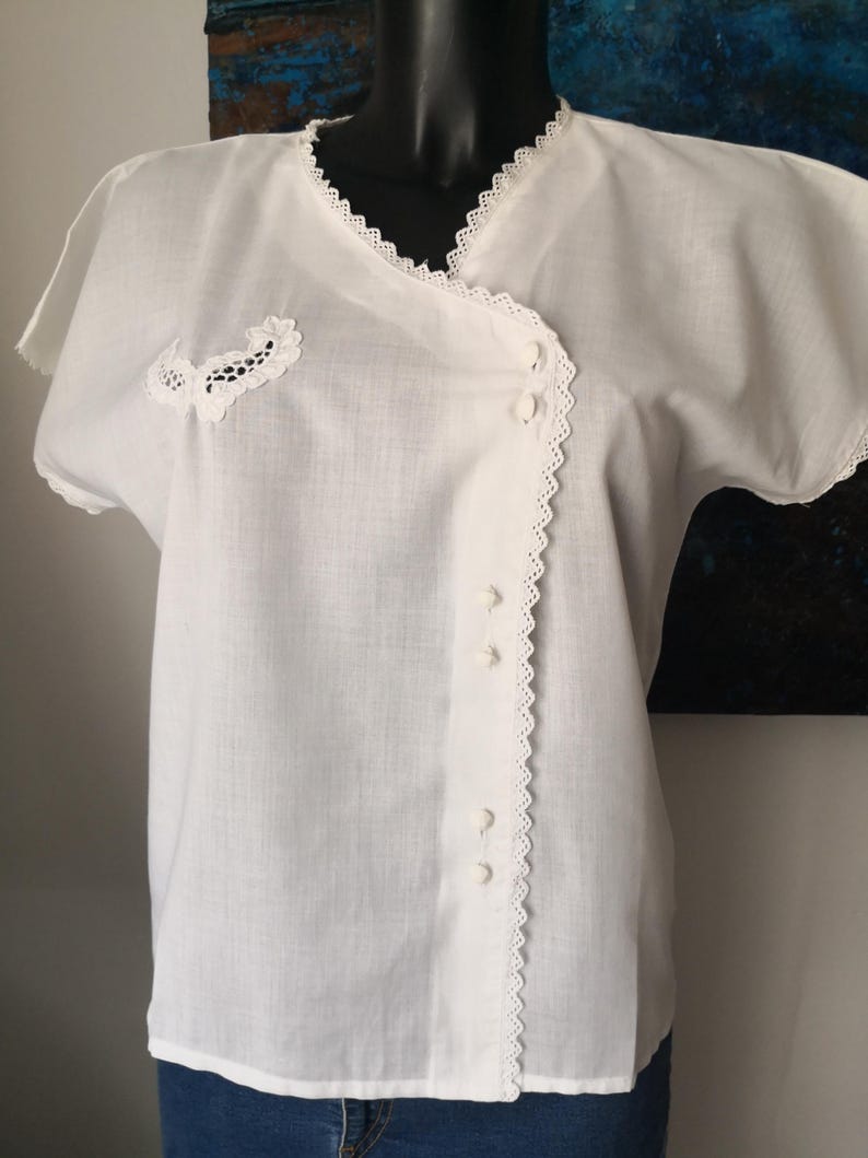 May include: White short-sleeved blouse with a wrap-style front. The blouse features delicate lace trim along the neckline, sleeves, and front closure. A small lace applique is on the left side. The blouse is buttoned down the front.
