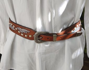 Puede incluir: Un cinturón de cuero marrón con hebilla plateada y costuras decorativas. El cinturón presenta diseños florales blancos y azules y se asegura con una hebilla plateada y un lazo de metal color latón.