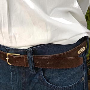 Può includere: Una cintura in pelle scamosciata marrone con una fibbia color oro e un accento color oro abbinato vicino al passante della cintura. La cintura è indossata con jeans blu e una camicia bianca.