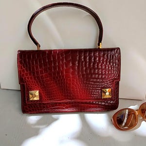 Peut inclure: Un sac à main rouge foncé avec un motif texturé en peau de crocodile, doté d'une anse arquée unique et de deux accents métalliques carrés dorés sur le rabat avant. Le sac présente une forme rectangulaire structurée et une finition brillante.