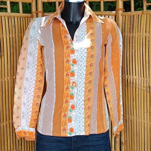 Puede incluir: Camisa vintage con botones y diseño patchwork. La camisa presenta rayas verticales en naranja, blanco y estampados florales. Se ven las mangas largas y el cuello. La camisa se exhibe en un maniquí.