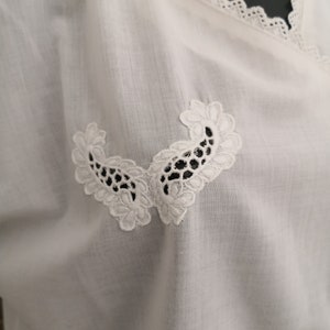 May include: White blouse with a V-neckline and delicate lace trim. The blouse features two symmetrical, embroidered floral appliques near the neckline. The fabric appears to be a lightweight, breathable material.
