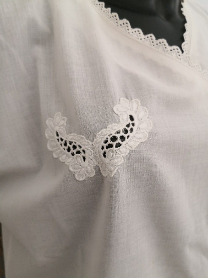 May include: White blouse with a V-neckline and delicate lace trim. The blouse features two symmetrical, embroidered floral appliques near the neckline. The fabric appears to be a lightweight, breathable material.