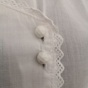 May include: Close-up of a white blouse with two faceted buttons. The blouse features a delicate lace trim along the neckline and button placket. The fabric appears to be a lightweight, breathable material.