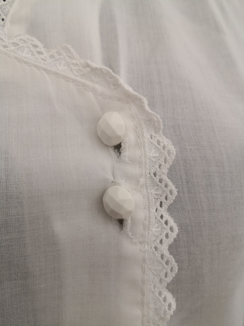 May include: Close-up of a white blouse with two faceted buttons. The blouse features a delicate lace trim along the neckline and button placket. The fabric appears to be a lightweight, breathable material.
