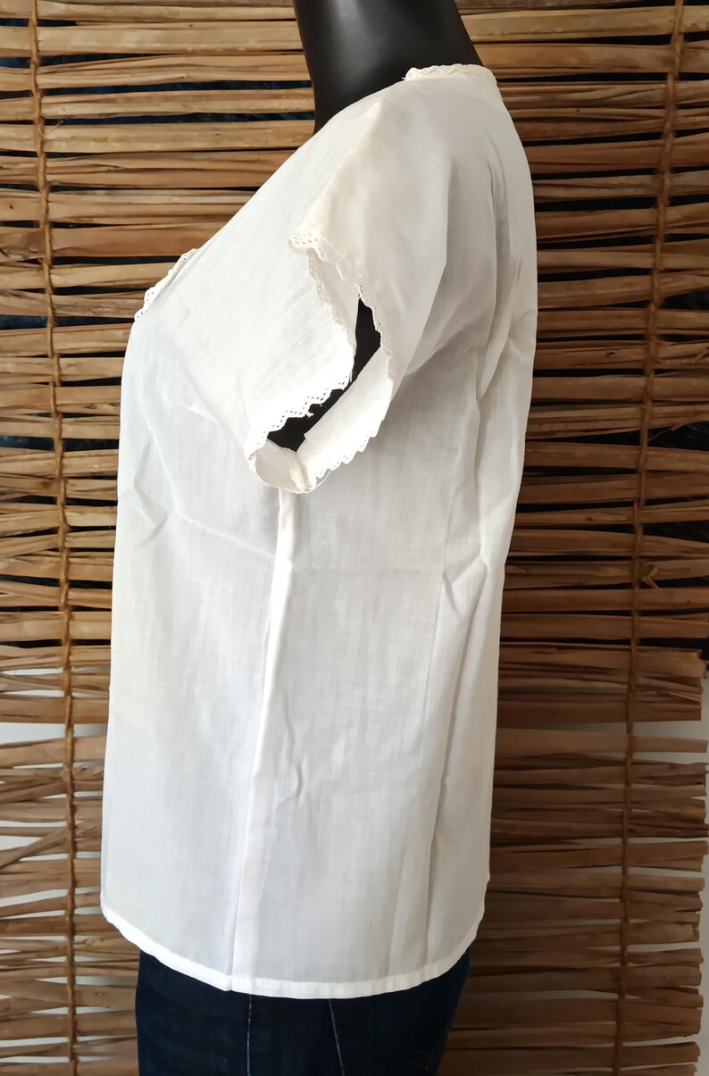 May include: A white, short-sleeved blouse with delicate lace trim along the neckline and sleeve edges. The blouse is made of a lightweight fabric and has a simple, classic design. The garment is displayed on a black mannequin.