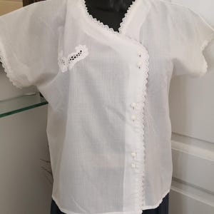 May include: A white, short-sleeved blouse with a wrap-style front. The blouse features delicate lace trim along the neckline, sleeves, and front closure. Small white buttons fasten the front. A small floral lace applique is on the left side.