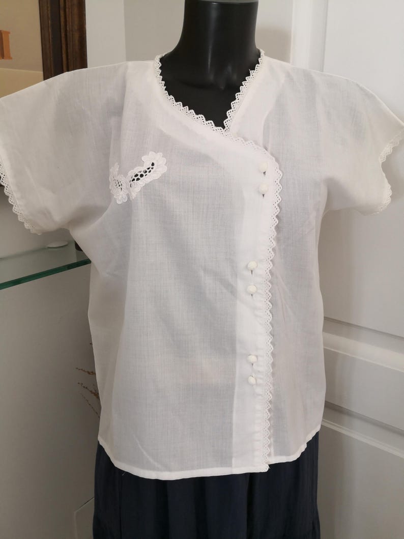 May include: A white, short-sleeved blouse with a wrap-style front. The blouse features delicate lace trim along the neckline, sleeves, and front closure. Small white buttons fasten the front. A small floral lace applique is on the left side.
