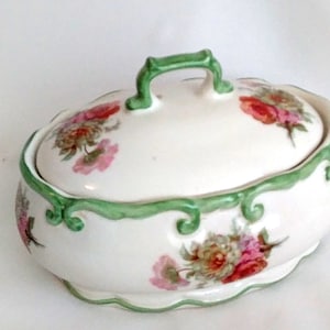 Op de afbeelding: Een witte keramische sieradendoos met een deksel, versierd met roze, rode en groene bloemmotieven. De doos heeft een geschulpte rand en een groene handgreep. De rand is groen.