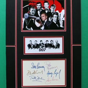 Könnte beinhalten: Ein gerahmter Druck mit Bildern von Schauspielern in Anzügen, einige halten Waffen. Der Druck enthält die Nummer "007" und mehrere Unterschriften. Der Rahmen ist schwarz mit roten Rändern. Das Gesamtthema ist ein Film.