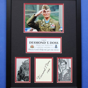 Puede incluir: Un memorial enmarcado en negro con el Cabo Desmond T. Doss, que incluye fotos, una firma y texto. La imagen superior muestra a Doss saludando con uniforme. El marco tiene un paspartú rojo, sobre un fondo azul.