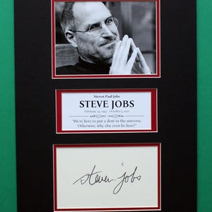 May include: A framed black and white portrait of Steve Jobs, with his name and signature below. The frame includes a quote: "We're here to put a dent in the universe. Otherwise, why else even be here?"