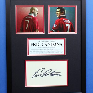 May include: A framed piece of memorabilia featuring Éric Cantona in a red Manchester United jersey with the number 7. The frame includes his name, team details, a quote, and his signature. The frame is black with white and red mounts.