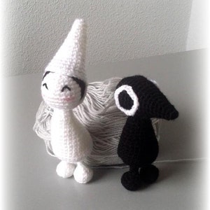May include: Two crocheted stuffed toys, one white with a pointed hat and the other black with a long beak. The toys are sitting on a grey surface with a ball of yarn.