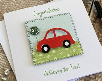 Puede incluir: Una tarjeta de felicitación blanca con el texto Congratulations y On Passing Your Test! en verde. Un recorte de papel cuadrado muestra un coche rojo sobre un campo floral verde con una pequeña señal verde de GO y costuras decorativas.