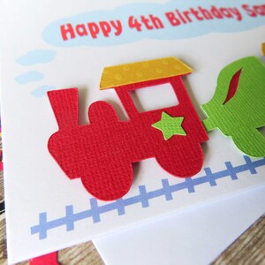 May include: A white birthday greeting card featuring a layered paper train design with a red engine, a yellow roof, a green star, and a green carriage. The text reads Happy 4th Birthday Sam inside a light blue cloud shape above a blue train track.