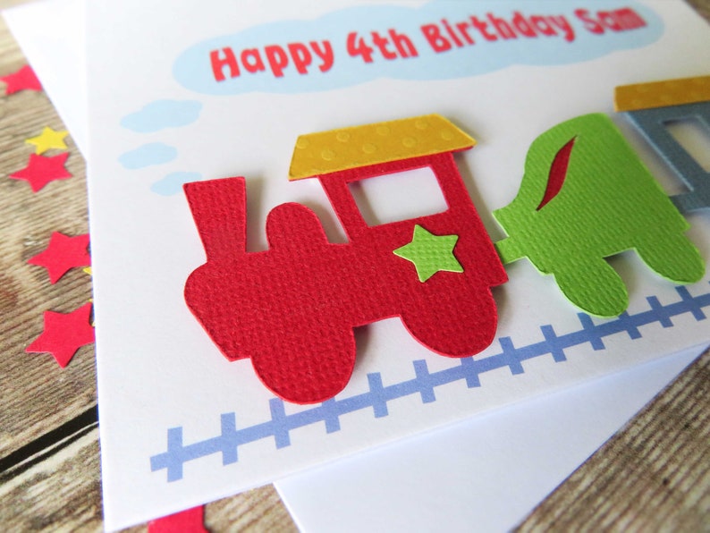 May include: A white birthday greeting card featuring a layered paper train design with a red engine, a yellow roof, a green star, and a green carriage. The text reads Happy 4th Birthday Sam inside a light blue cloud shape above a blue train track.