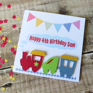 May include: A square white greeting card featuring a colorful bunting banner at the top and the text Happy 4th Birthday Sam in red letters inside a light blue cloud. A red, green, and blue train sits on a blue track at the bottom of the card.